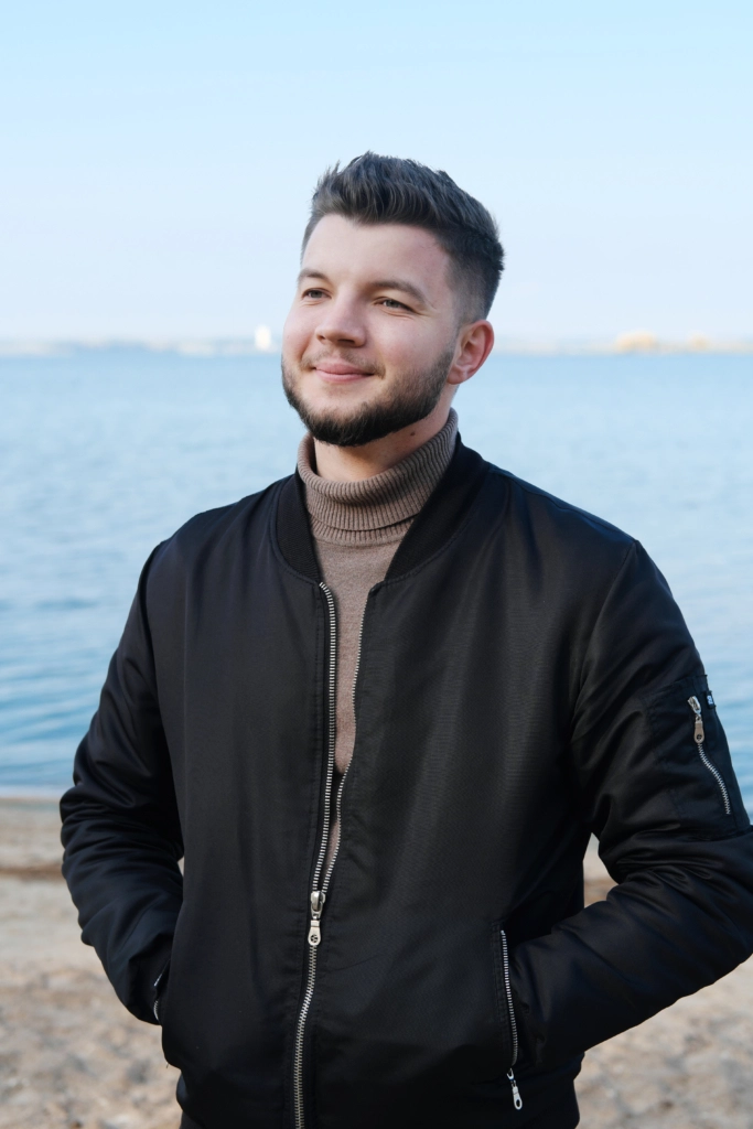 A young man in a black jacket is standing on the beach against the backdrop of a lake and smiling.