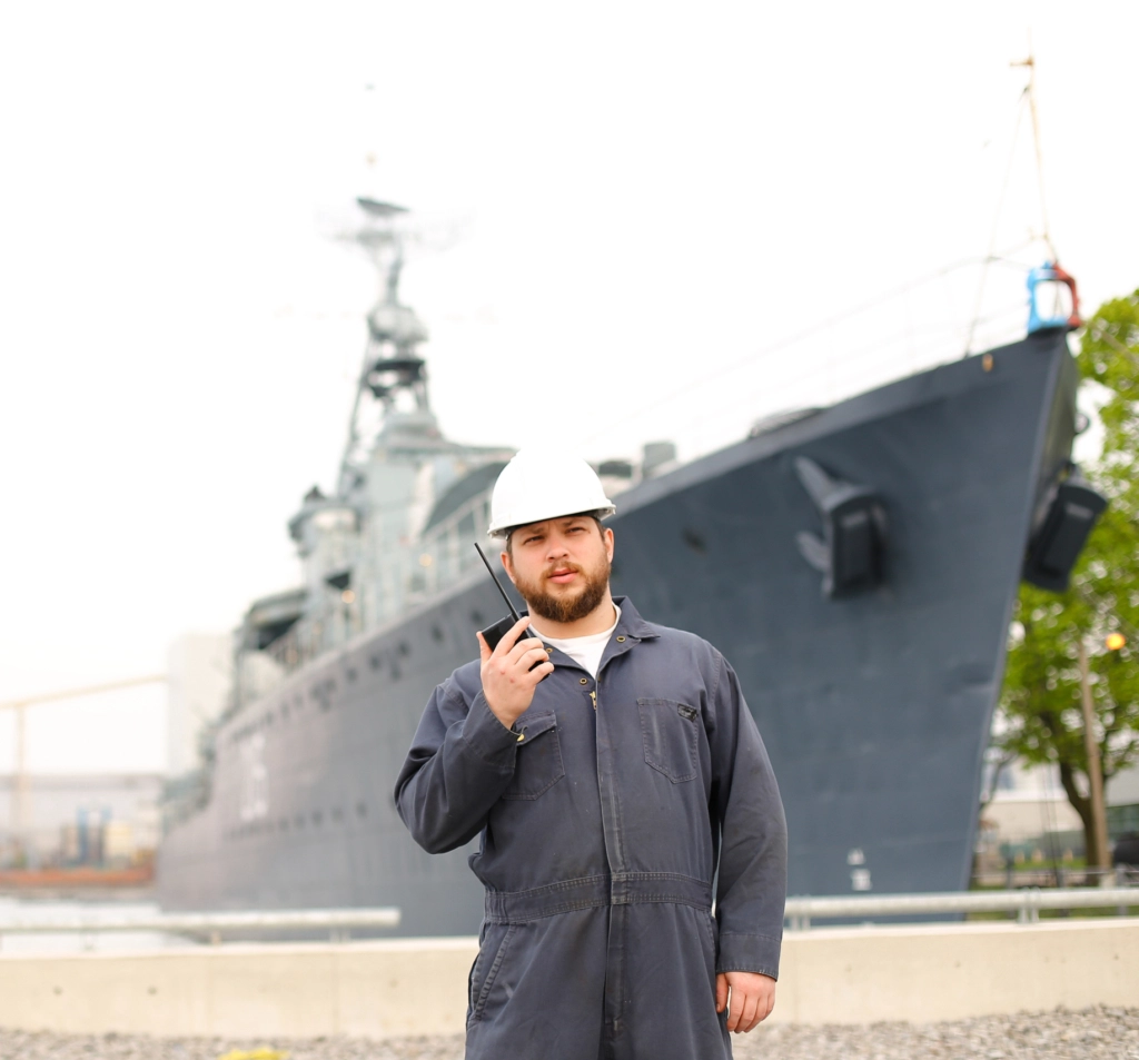 Marine deck officer talking by walkie talkie radio and standing near offshore vessel in bakground.